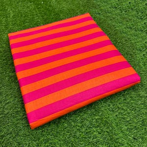 Puede incluir: Una tela doblada con rayas horizontales alternas de color naranja brillante y rosa intenso. La tela está sobre un lecho de hierba verde.