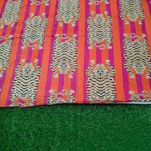 Può includere: Un tessuto vibrante con un motivo ripetuto di tigri stilizzate con strisce nere e pelliccia color tan. Le tigri sono poste su uno sfondo di strisce verticali in rosa brillante e arancione. Il tessuto è probabilmente cotone o un materiale simile.