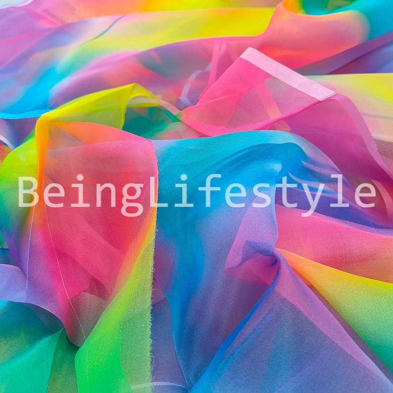May include: A vibrant, multi-colored fabric with a rainbow gradient of pink, yellow, blue, and green hues. The sheer material is folded and draped, creating a dynamic display. The text "BeingLifestyle" is visible in white.