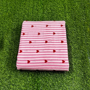 Könnte beinhalten: Gefalteter Stoff mit rosa-weißem Streifenmuster, verziert mit roten Herzmotiven. Das Material scheint Baumwolle oder ein ähnliches Textil zu sein, ideal zum Basteln oder Nähen. Das Muster ist schlicht und ansprechend.