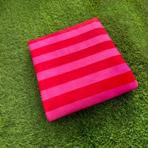 Puede incluir: Una pieza de tela cuadrada que presenta un patrón repetitivo de rayas horizontales en tonos alternos de rosa brillante y rojo vibrante. El tejido está doblado en una forma plana y ordenada, mostrando un tejido consistente y color uniforme.
