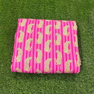 Op de afbeelding: Een gevouwen stof met een roze en witte gestreepte achtergrond. De stof is bedekt met een herhalend patroon van gele cheeta's met zwarte vlekken. Geschikt voor kleding of woondecoratie.