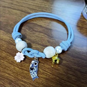May include: A light blue bracelet with a knotted design, featuring wooden beads, white pearls, and a gold bell charm. A blue and white carp pendant and a white flower charm add detail to this unique accessory.