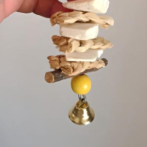 May include: A bird toy with a pink bead, white blocks, woven raffia, a yellow bead, and a small gold bell. The toy is suspended from a metal loop.
