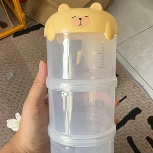 Puede incluir: Un dispensador de fórmula transparente de tres niveles con una tapa amarilla en forma de oso. El dispensador tiene marcas de medición en mililitros. La tapa presenta una cara de oso de dibujos animados con los ojos cerrados.