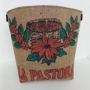 Puede incluir: Una cesta de yute con un diseño floral. La cesta presenta una flor de pascua roja, hojas verdes y bayas rojas. Las palabras "LA PASTORA" están impresas en rojo en la parte delantera. La cesta tiene una parte superior curva y asas negras.