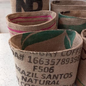 May include: Several burlap storage baskets with varying designs. Some have pink or green stripes along the top edge. One basket displays the word "ND" in black. Another has text that reads "BRAZIL SANTOS NATURAL COFFEE".