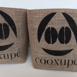 Puede incluir: Dos recipientes de yute beige con un diseño de granos de café negros y la palabra "cooxupé" impresa. Los recipientes tienen una textura rugosa y un diseño de cara sonriente.