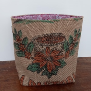 Peut inclure: Un panier en toile de jute beige avec un motif floral. Le motif comprend une illustration de grains de café, un poinsettia et des feuilles vertes. L'intérieur est doublé d'un tissu floral rose. Le panier est posé sur une surface en bois.