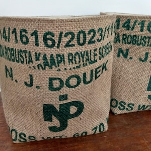 May include: Two burlap planters with green text and numbers. The text includes "ROBUSTA KAAPI ROYALE SCREEN" and "N. J. DOUEK". The planters are cylindrical and sit on a wooden surface.