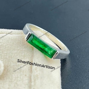 May include: A silver ring with a rectangular emerald green gemstone set in a bezel setting.