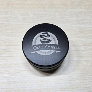 May include: A black coffee grinder with the text "OMG Coffee®" and a coffee cup logo. The grinder is round and made of metal. The logo is etched into the top of the grinder.
