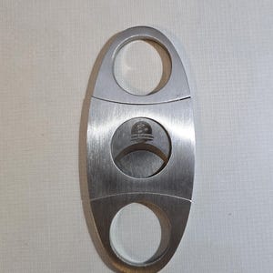 May include: A silver-colored metal cigar cutter with an oval shape and two finger holes. The center features a circular blade mechanism with the text Chic Lighters printed in black on the metal surface between the two circular openings.