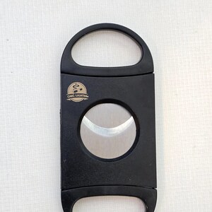 May include: A black cigar cutter with a dual-blade guillotine mechanism and two finger loops. The front features a circular logo with the text OMG LIGHTER and a graphic of a cigar with smoke. The blades are metallic silver and circular in shape.