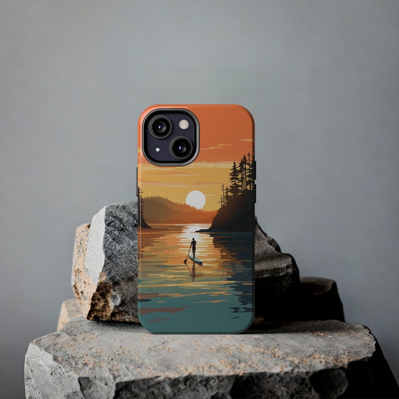 May include: A phone case with a sunset scene featuring a silhouette of a person paddleboarding on a lake. The case has a dark blue, teal, and orange color scheme.