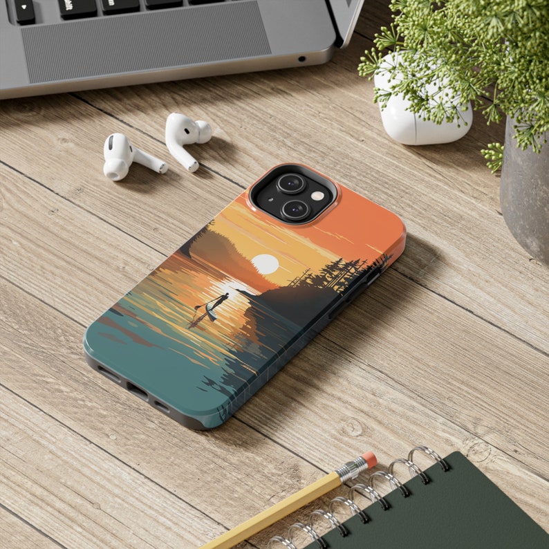 May include: A phone case with a sunset scene featuring a person paddleboarding on a lake. The case is made of a hard plastic material and has a matte finish. The design is printed directly onto the case and features a vibrant color palette.