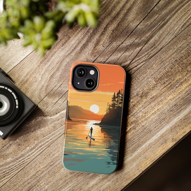 May include: A phone case with a sunset scene featuring a person paddleboarding on a lake. The case has a silhouette of trees and a bright orange sunset in the background.