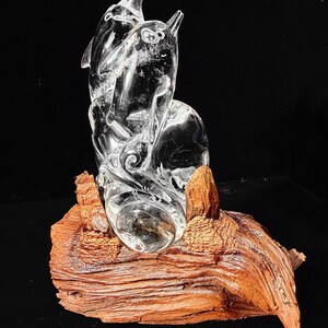 Pode incluir: Uma escultura de cristal transparente de dois golfinhos saltando de uma onda, montada em um peda&ccedil;o de madeira natural, marrom avermelhado. A escultura &eacute; detalhada e transparente, com o texto "Crystal Patronum Shop" na parte inferior.