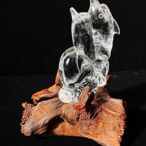 Pode incluir: Uma escultura de cristal transparente de uma figura animal estilizada, possivelmente um c&atilde;o ou um lobo, montada sobre um peda&ccedil;o de madeira natural castanho-avermelhada. A escultura tem um aspeto liso e polido.