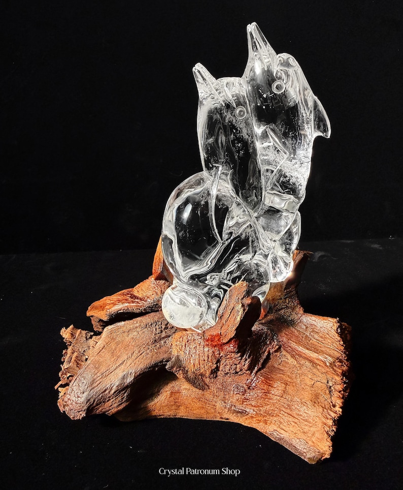 Pode incluir: Uma escultura de cristal transparente de uma figura animal estilizada, possivelmente um c&atilde;o ou um lobo, montada sobre um peda&ccedil;o de madeira natural castanho-avermelhada. A escultura tem um aspeto liso e polido.