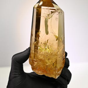 Puede incluir: Una punta de cristal de citrino pulido con forma hexagonal, que presenta un tono amarillo dorado translúcido, fracturas internas e inclusiones visibles. La base es rugosa y texturizada, mientras que la parte superior termina en punta.