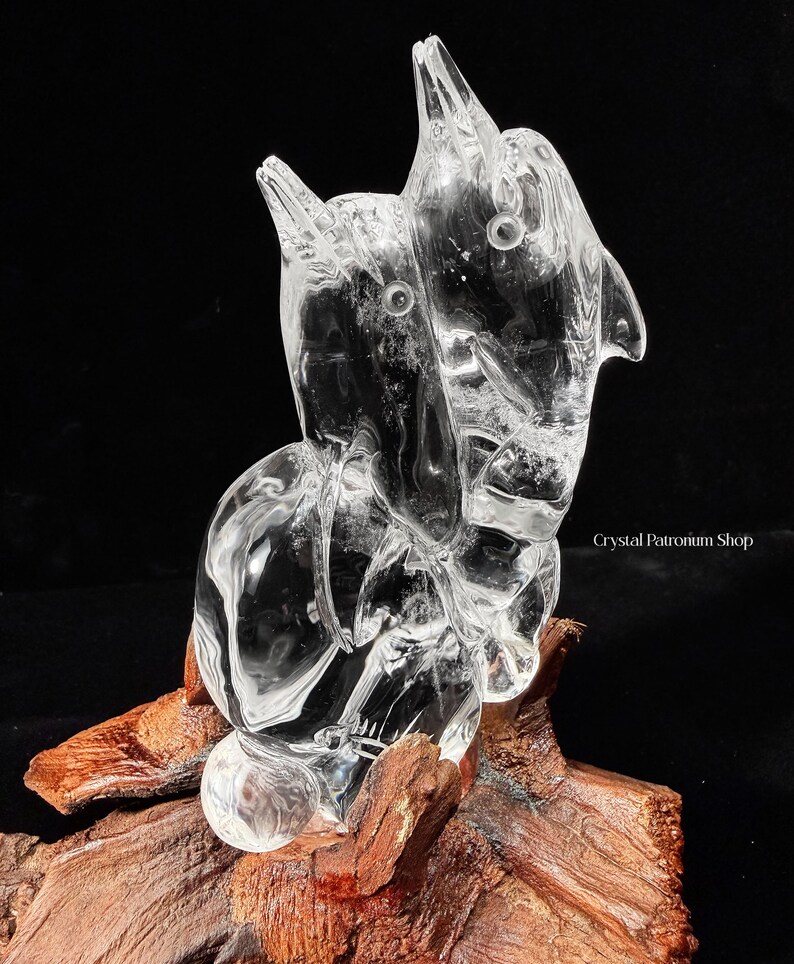 Pode incluir: Uma escultura de cristal transparente de dois golfinhos, montada sobre um peda&ccedil;o de madeira. Os golfinhos s&atilde;o esculpidos de forma intrincada com detalhes vis&iacute;veis. A escultura est&aacute; sobre um fundo preto, destacando a clareza do cristal.