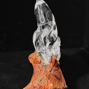 Pode incluir: Uma escultura de vidro transparente, com detalhes internos, montada sobre uma base de madeira natural castanho-avermelhada. O vidro tem uma superf&iacute;cie lisa e polida, enquanto a base tem uma apar&ecirc;ncia &aacute;spera e texturizada. O texto "Crystal Patronum Shop" est&aacute; na parte inferior.