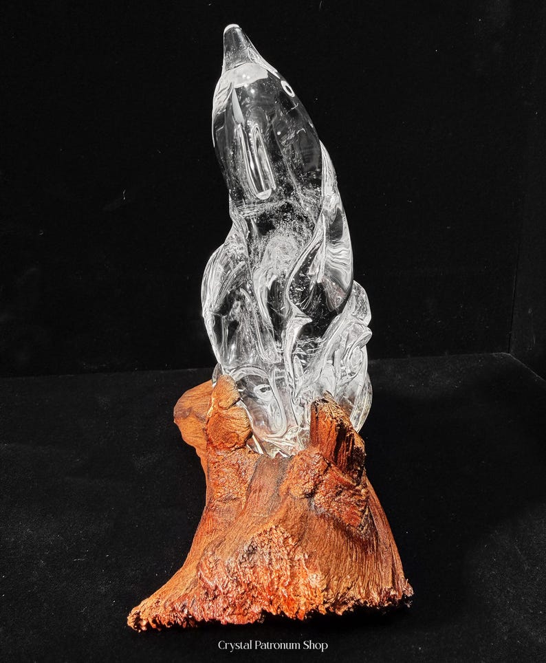 Pode incluir: Uma escultura de vidro transparente, com detalhes internos, montada sobre uma base de madeira natural castanho-avermelhada. O vidro tem uma superf&iacute;cie lisa e polida, enquanto a base tem uma apar&ecirc;ncia &aacute;spera e texturizada. O texto "Crystal Patronum Shop" est&aacute; na parte inferior.