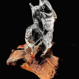 Pode incluir: Uma escultura de vidro transparente de dois golfinhos saltando para cima, montada em um peda&ccedil;o de madeira flutuante. Os golfinhos s&atilde;o detalhados com barbatanas e olhos, e a madeira tem uma apar&ecirc;ncia natural e texturizada. A escultura est&aacute; em um fundo preto.