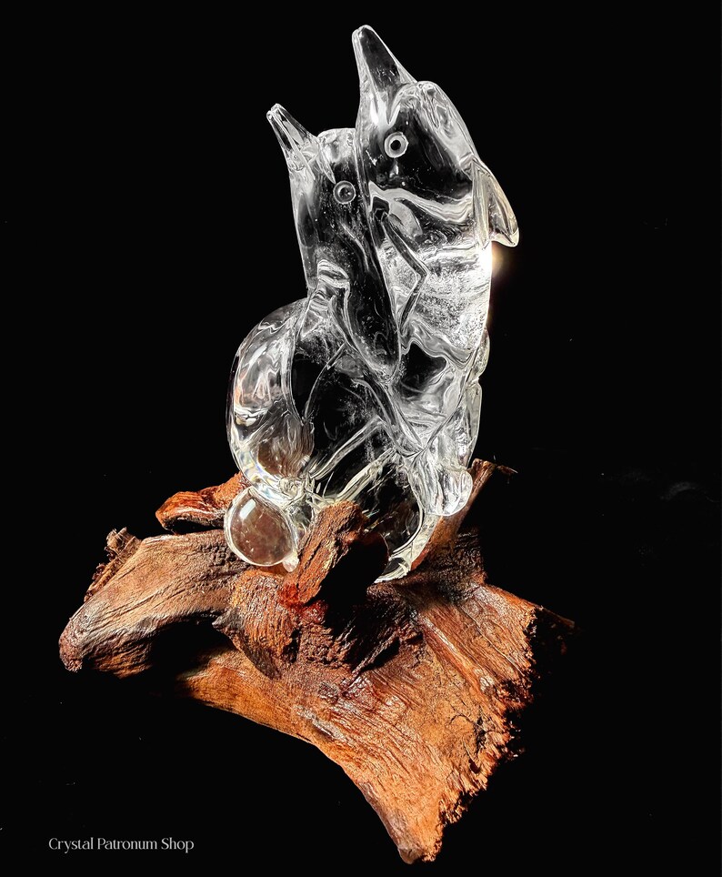 Pode incluir: Uma escultura de vidro transparente de dois golfinhos saltando para cima, montada em um peda&ccedil;o de madeira flutuante. Os golfinhos s&atilde;o detalhados com barbatanas e olhos, e a madeira tem uma apar&ecirc;ncia natural e texturizada. A escultura est&aacute; em um fundo preto.