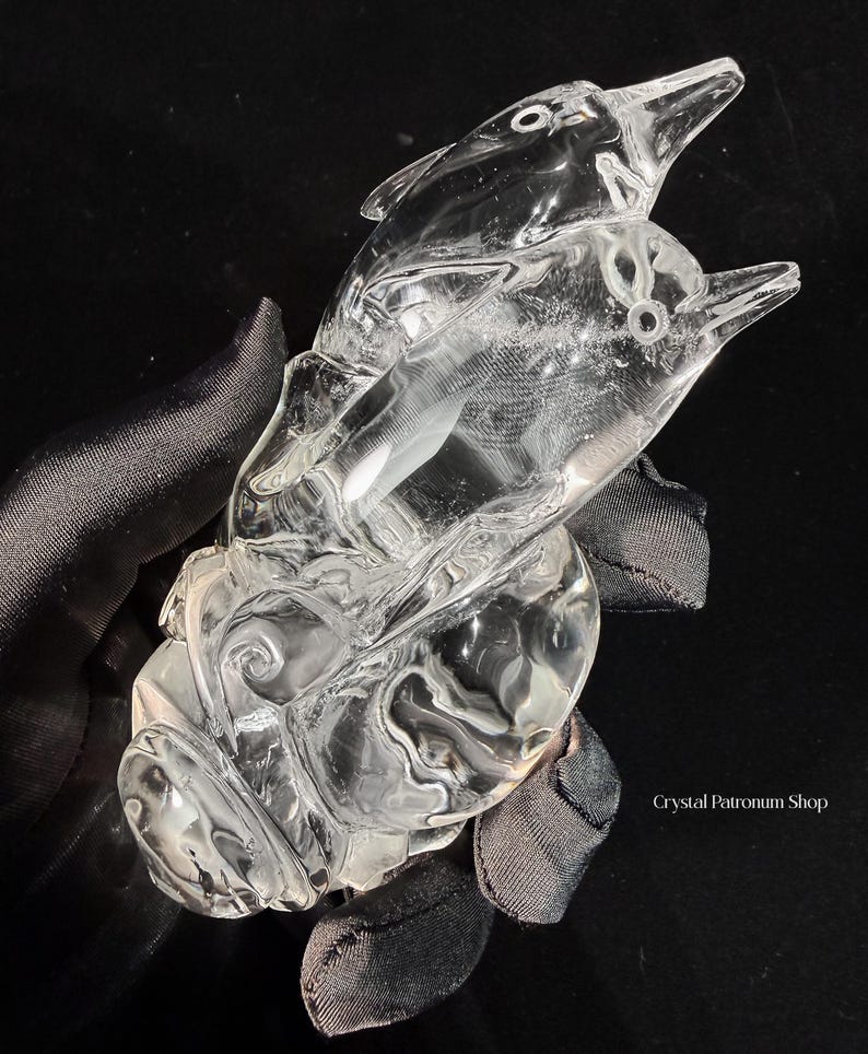 Pode incluir: Uma escultura de cristal transparente de dois golfinhos saltando da &aacute;gua. Os golfinhos s&atilde;o esculpidos de forma intrincada com detalhes, e a escultura assenta sobre uma base com padr&otilde;es de ondas rodopiantes. O cristal &eacute; transparente.