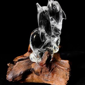 Pode incluir: Uma escultura de cristal transparente de dois golfinhos estilizados, montada sobre um peda&ccedil;o de madeira flutuante natural marrom escuro. Os golfinhos s&atilde;o esculpidos de forma intrincada com formas lisas e arredondadas e barbatanas pontiagudas. O conjunto est&aacute; sobre fundo preto.