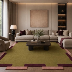 May include: A modern living room featuring a large sectional sofa, a coffee table, and a patterned area rug. The rug has a green center with burgundy and pink geometric borders. The room is decorated in neutral tones with a large window and a decorative lamp.