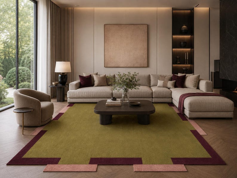 May include: A modern living room featuring a large sectional sofa, a coffee table, and a patterned area rug. The rug has a green center with burgundy and pink geometric borders. The room is decorated in neutral tones with a large window and a decorative lamp.
