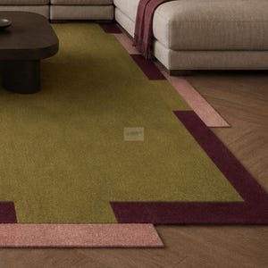 May include: A modern area rug with a geometric design. The rug features a central olive green field, bordered by a deep burgundy and a blush pink accent. The rug is placed on a wooden floor, with a section of a sofa visible in the background.