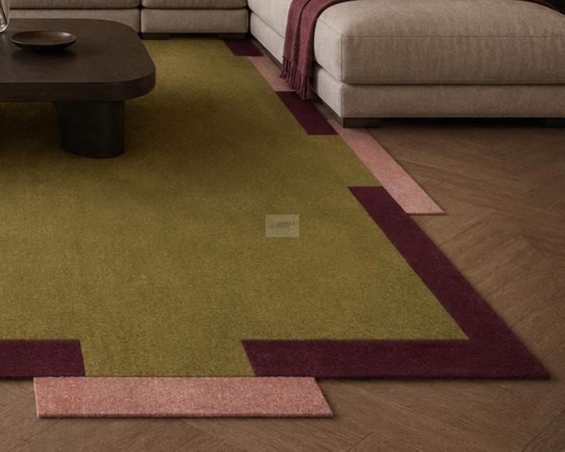 May include: A modern area rug with a geometric design. The rug features a central olive green field, bordered by a deep burgundy and a blush pink accent. The rug is placed on a wooden floor, with a section of a sofa visible in the background.