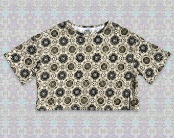 Puede incluir: Una camiseta corta de manga corta con un patrón geométrico en negro y dorado sobre un fondo crema.