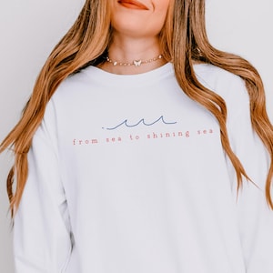 May include: White long-sleeve shirt with the text "from sea to shining sea" in red and blue. Above the text is a blue wave design. The shirt is a comfortable, casual style.