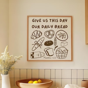 Op de afbeelding: Een ingelijste print met de tekst "GIVE US THIS DAY OUR DAILY BREAD" en illustraties van verschillende broodsoorten. De print zit in een lichtbruine lijst en wordt in een keuken weergegeven.