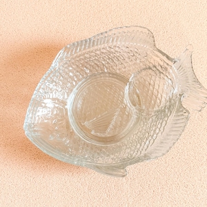 Könnte beinhalten: Klarer Servierteller aus Glas in Form eines Fisches. Die Schale hat ein großes zentrales Fach und ein kleineres, kreisförmiges Fach. Das Fischdesign umfasst Schuppen, Flossen und einen Schwanz.