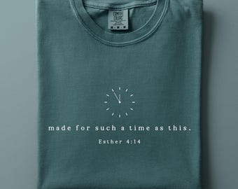 May include: A teal crew neck t-shirt featuring a white graphic of a clock face with the text "made for such a time as this." and "Esther 4:14" printed below it. The shirt has a Comfort Colors brand label visible at the inside of the neckline.