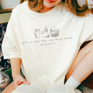 May include: White t-shirt featuring a graphic of a jar, a sack of flour, and a basket of bread. The text "give us this day our daily bread. matthew 6:11" is printed below the graphic.