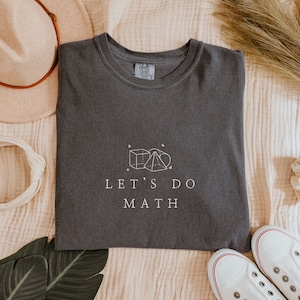 May include: A dark gray t-shirt with a white graphic of geometric shapes and the text "LET'S DO MATH".