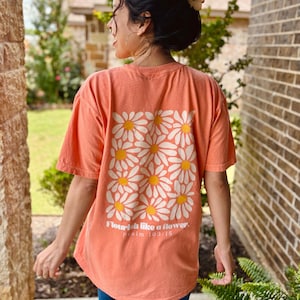 Könnte beinhalten: Ein korallenfarbenes Kurzarm-T-Shirt mit einer großen quadratischen Grafik auf der Rückseite, die aus weißen Gänseblümchen mit gelben Mitten besteht. Unter dem Blumenmuster steht der Text Flourish like a flower. psalm 103:15 in weißer Schrift.