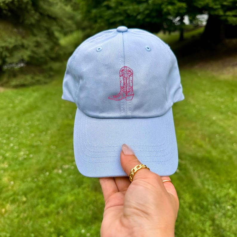 Pu&ograve; includere: Un berretto da baseball blu chiaro con un disegno di stivale da cowboy ricamato in rosa.