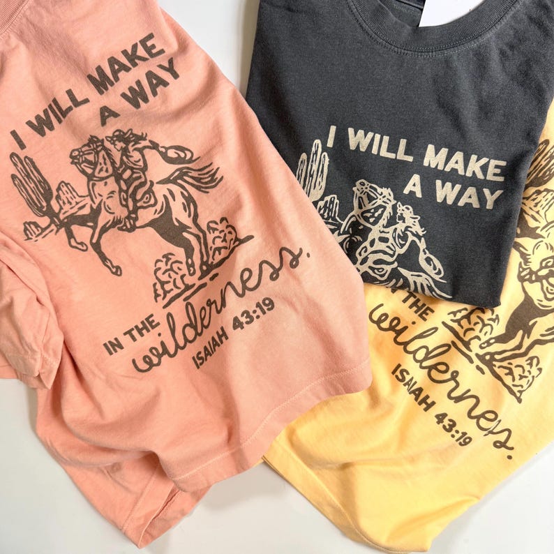 May include: Three t-shirts in peach, charcoal, and yellow, each featuring a graphic of a cowboy on a horse with cacti and the text "I WILL MAKE A WAY IN THE wilderness. ISAIAH 43:19". The shirts are made of soft fabric.