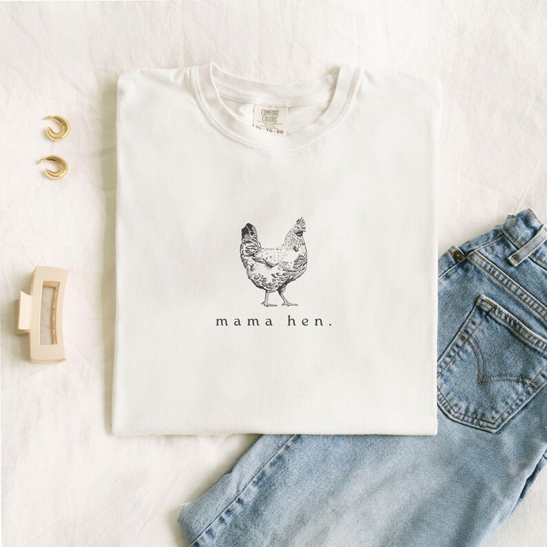 May include: White t-shirt with a black and white illustration of a hen and the text "mama hen." The shirt is paired with blue jeans, gold hoop earrings, and a cream-colored hair clip.