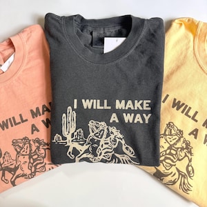 May include: Three folded t-shirts in peach, charcoal, and yellow. Each shirt features the text "I WILL MAKE A WAY" with a desert scene graphic. The shirts are made of a soft material and have a vintage look.