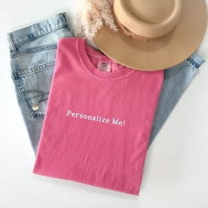 May include: A pink t-shirt with the text "Personalize Me!" in white. The shirt is folded and laid on a white surface with blue jeans and a brown hat.