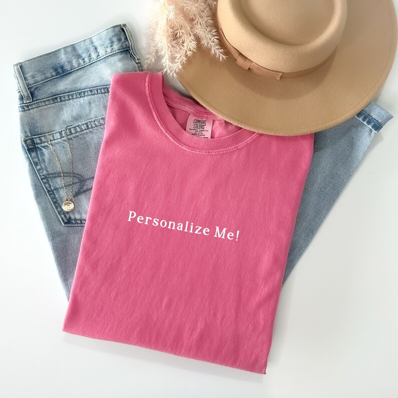 May include: A pink t-shirt with the text "Personalize Me!" in white. The shirt is folded and laid on a white surface with blue jeans and a brown hat.